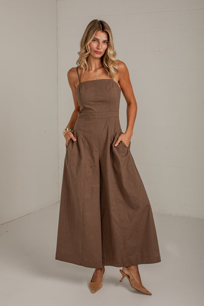 A woman modeling a brown wide-leg jumpsuit with thin straps, a square neckline, and side pockets, standing in a studio setting.