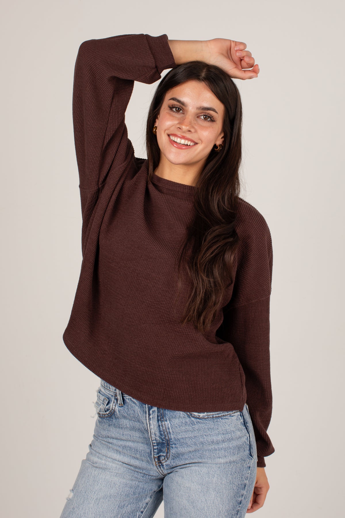 A woman wearing a dark brown long-sleeve knit sweater and light blue jeans, posing against a solid light gray background.