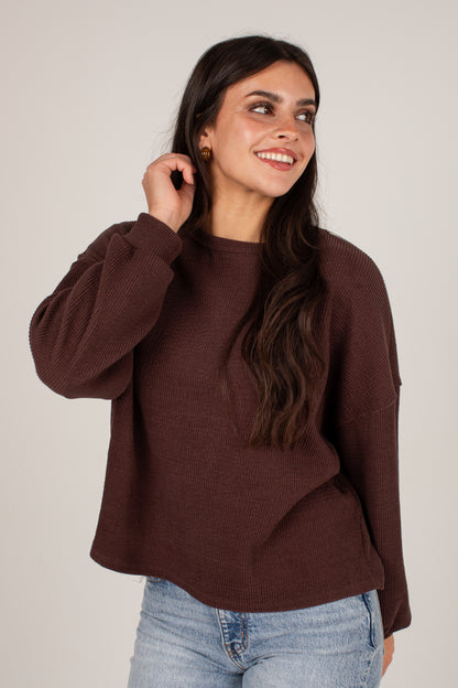 A woman wearing a dark brown ribbed knit sweater and light blue jeans, posing against a neutral light gray background.