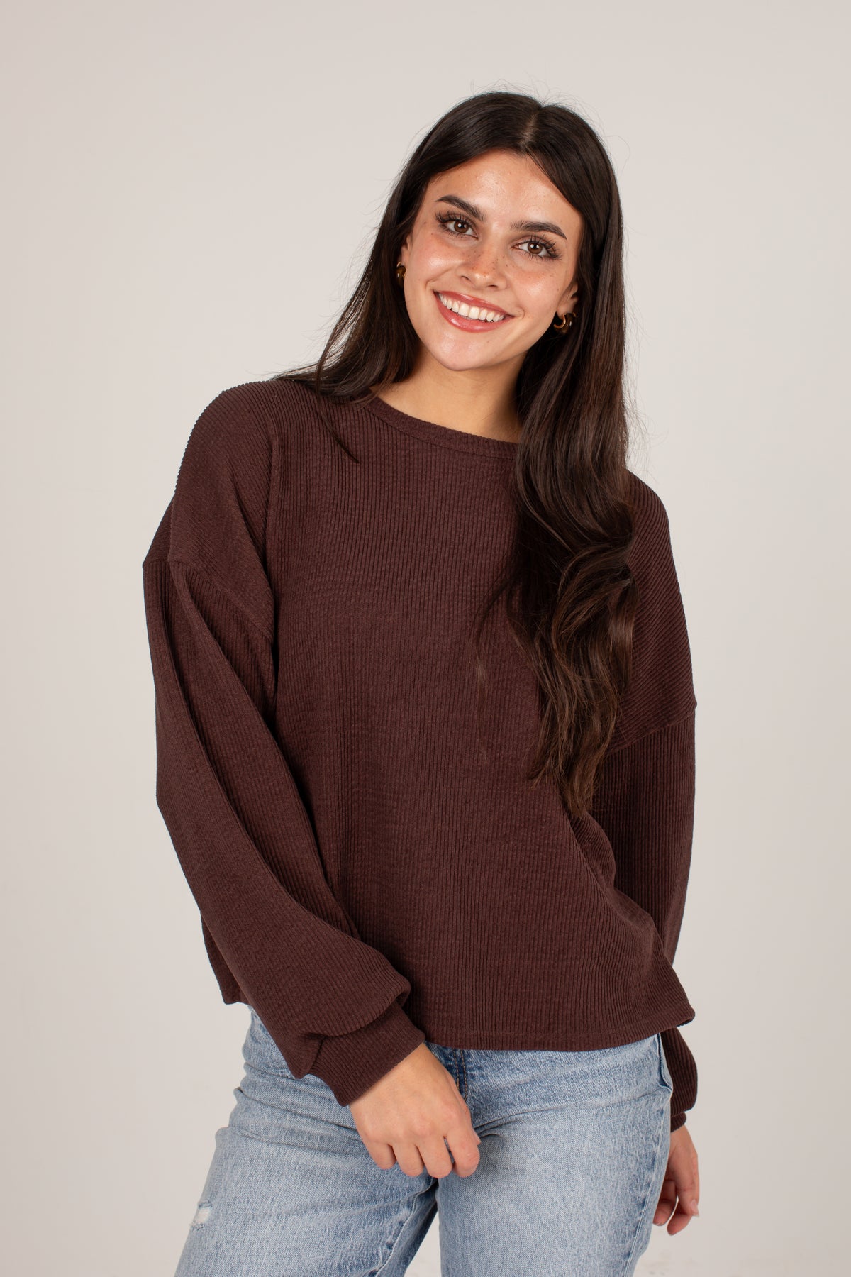 A woman with long dark hair wearing a dark brown waffle-knit crewneck sweatshirt and light blue jeans, standing against a plain light gray studio background.