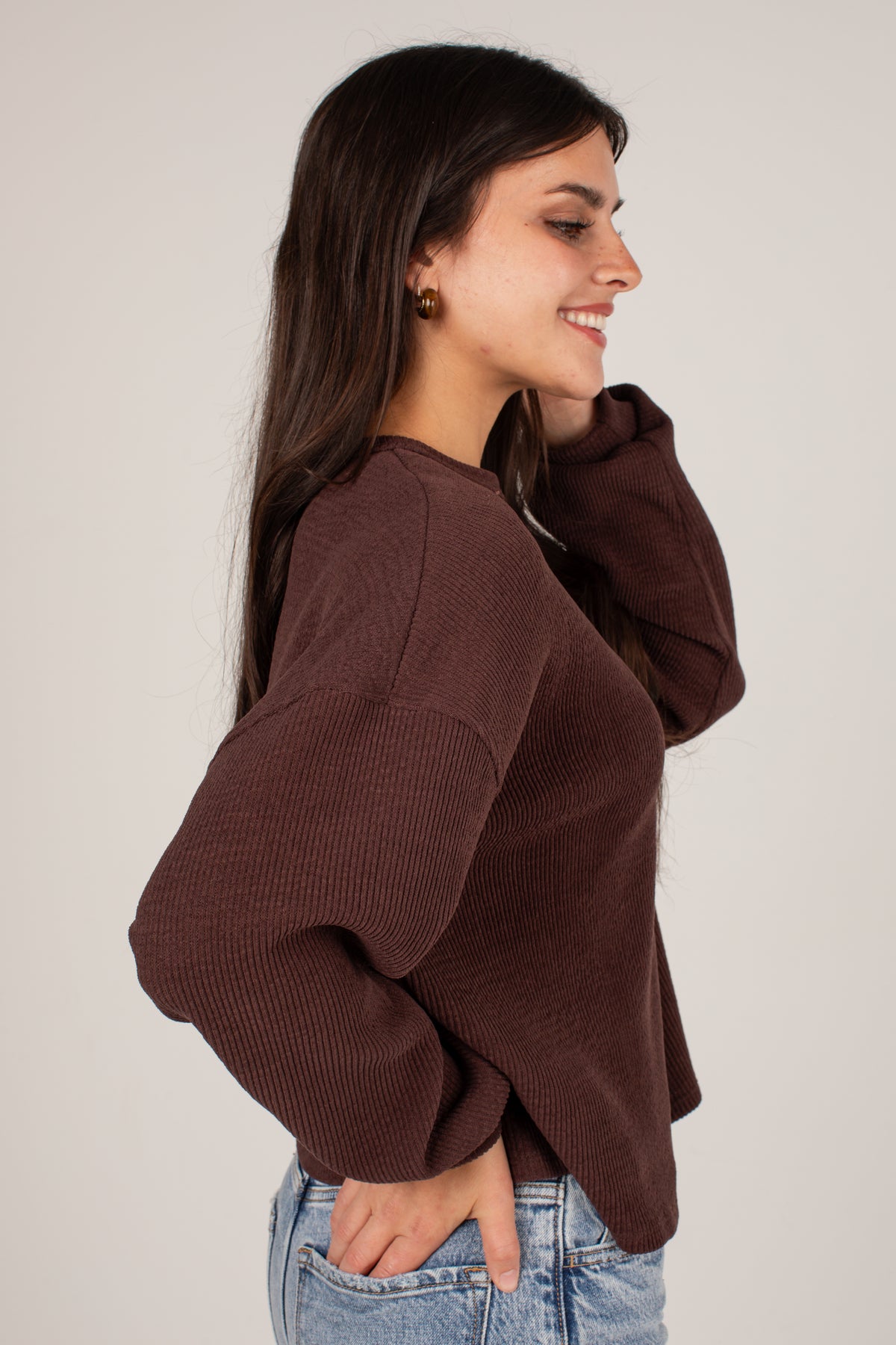 A woman wearing a dark brown ribbed knit sweater with balloon sleeves, paired with light blue jeans, shown from a side profile against a plain light gray background.