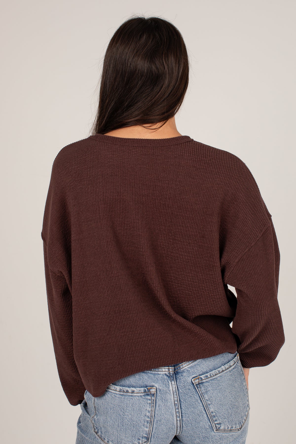 A woman viewed from behind wearing a dark brown waffle-knit long-sleeve sweater and light blue jeans against a plain light gray background.