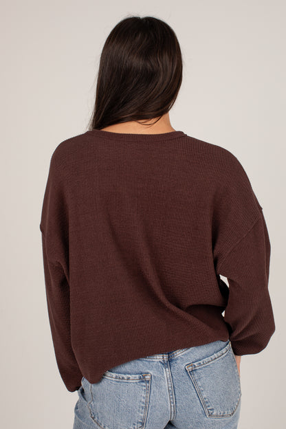 A woman viewed from behind wearing a dark brown waffle-knit long-sleeve sweater and light blue jeans against a plain light gray background.