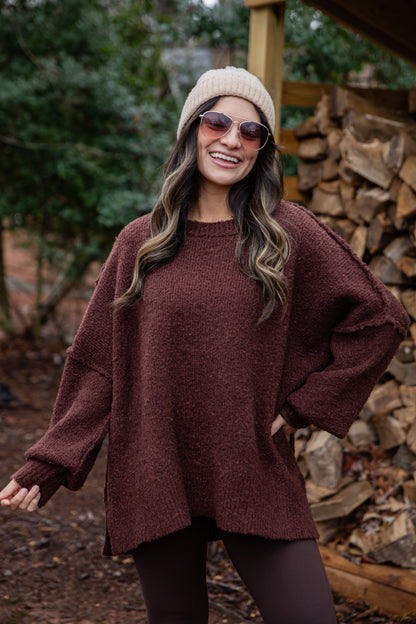 Woman wearing a maroon sweater and beige beanie outdoors with stacked firewood in the background