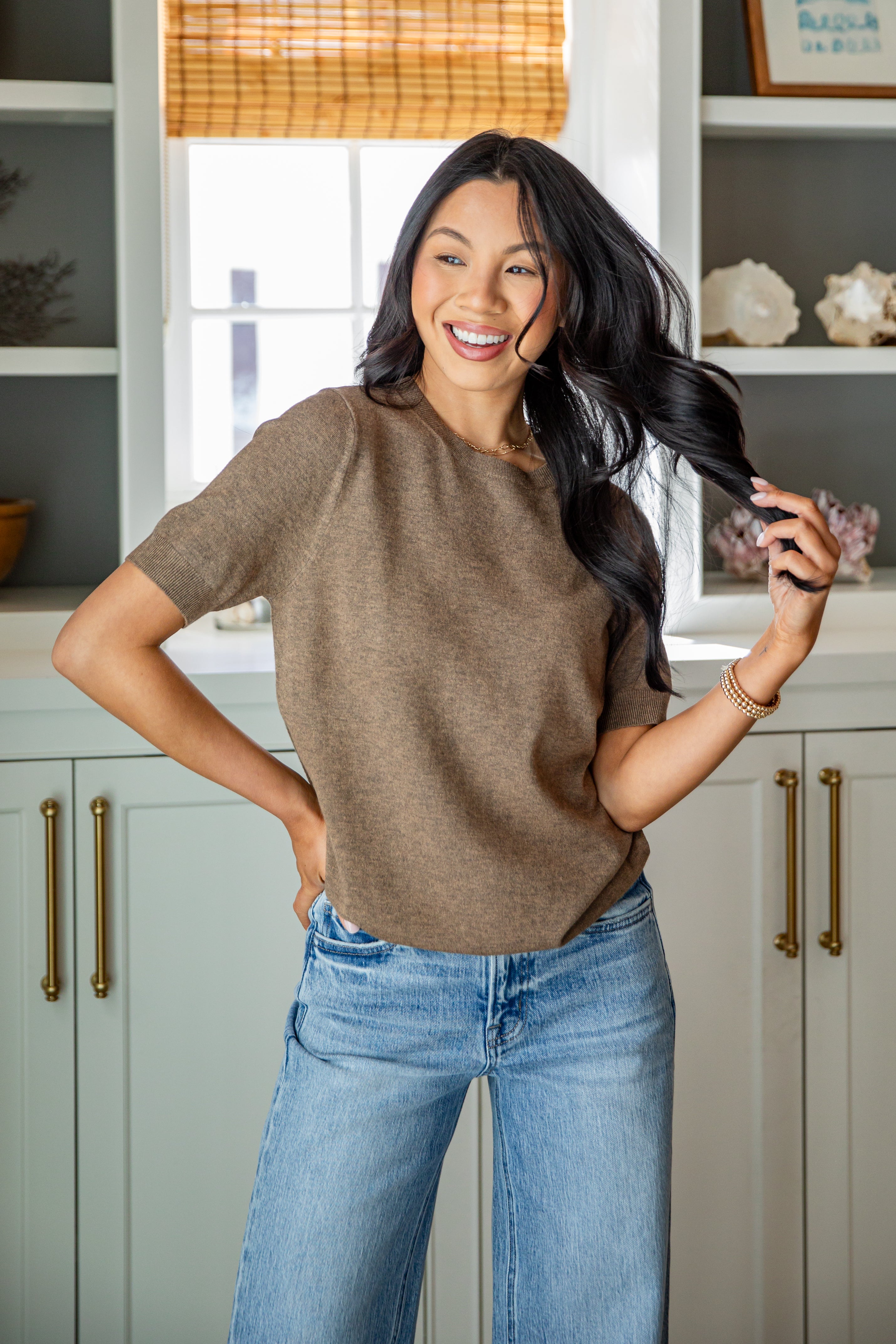 Woman in a brown sweater and blue jeans standing in a kitchen.