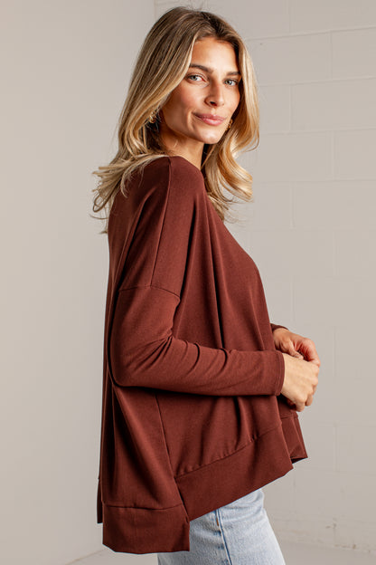 A woman wearing a rust-brown long-sleeve knit top with a relaxed, draped silhouette, paired with light-wash jeans, standing against a light gray brick wall.