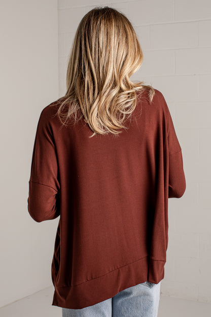 A woman viewed from behind wearing an oversized rust-brown long-sleeve top and light blue jeans, standing against a white brick wall.
