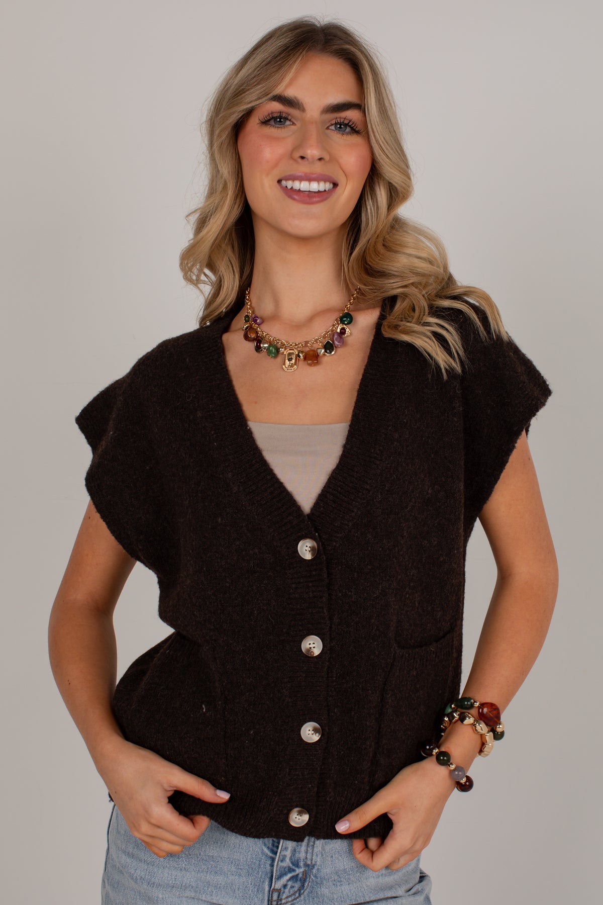 A smiling blonde woman wearing a dark brown sleeveless button-front vest over a beige top and light blue jeans, photographed against a plain light gray studio background.