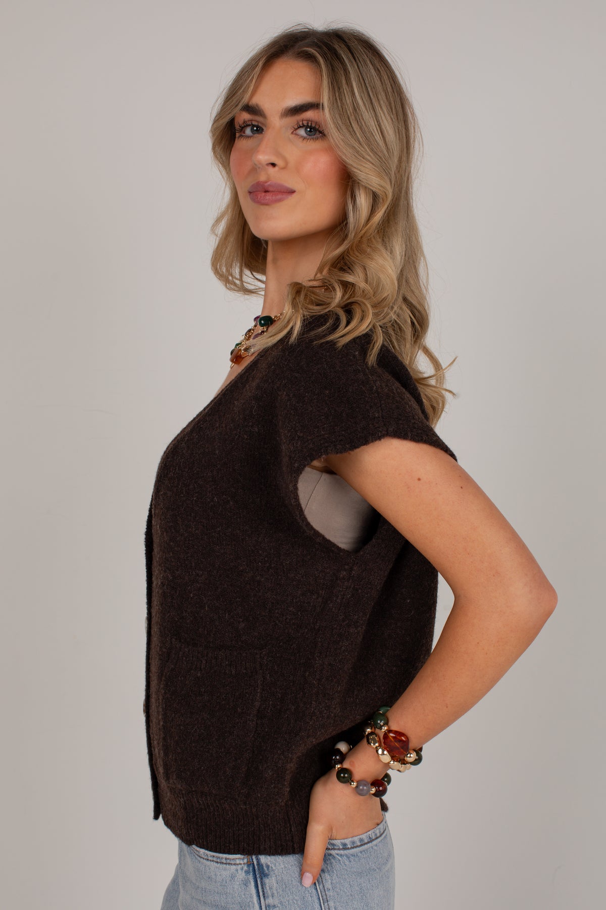A woman modeling a dark brown short-sleeve knit sweater vest over a light gray top, paired with light blue jeans, against a solid light gray background.