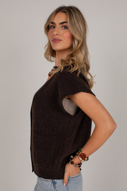 A woman modeling a dark brown short-sleeve knit sweater vest over a light gray top, paired with light blue jeans, against a solid light gray background.