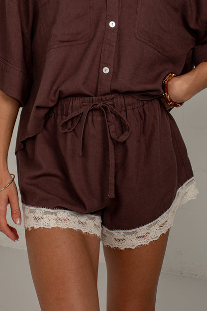 A close-up of a person wearing dark brown drawstring shorts with white lace trim at the hem, paired with a matching brown button-front top.