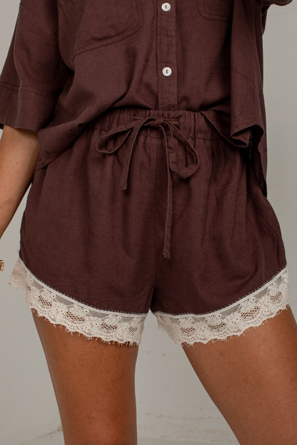 A close-up of a person wearing a dark brown two-piece outfit with matching shorts featuring a cream lace hem and a drawstring waist, paired with a matching short-sleeve button-up top.
