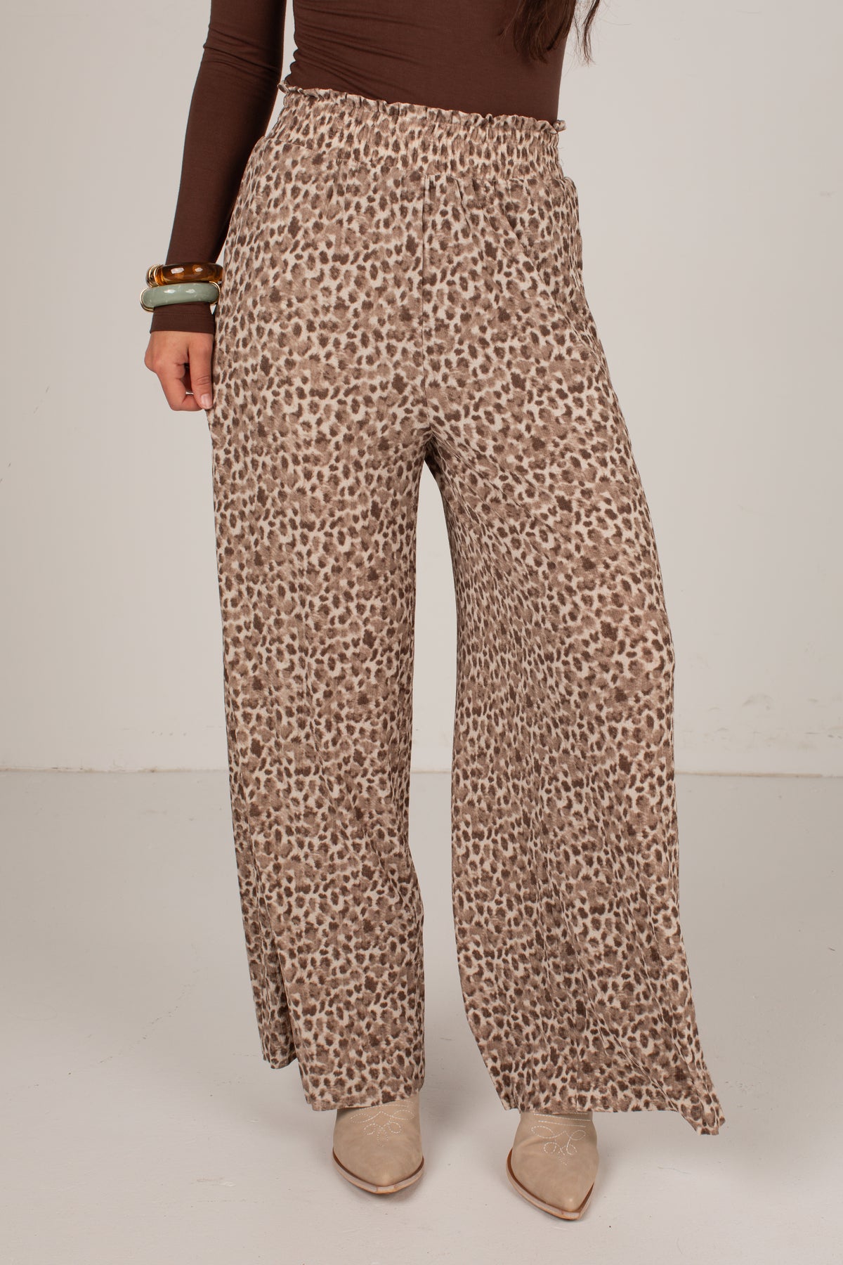A person wearing high-waisted, wide-leg pants with an allover leopard print pattern in brown and beige, paired with a brown long-sleeve top and beige pointed-toe heels against a plain light gray studio background.