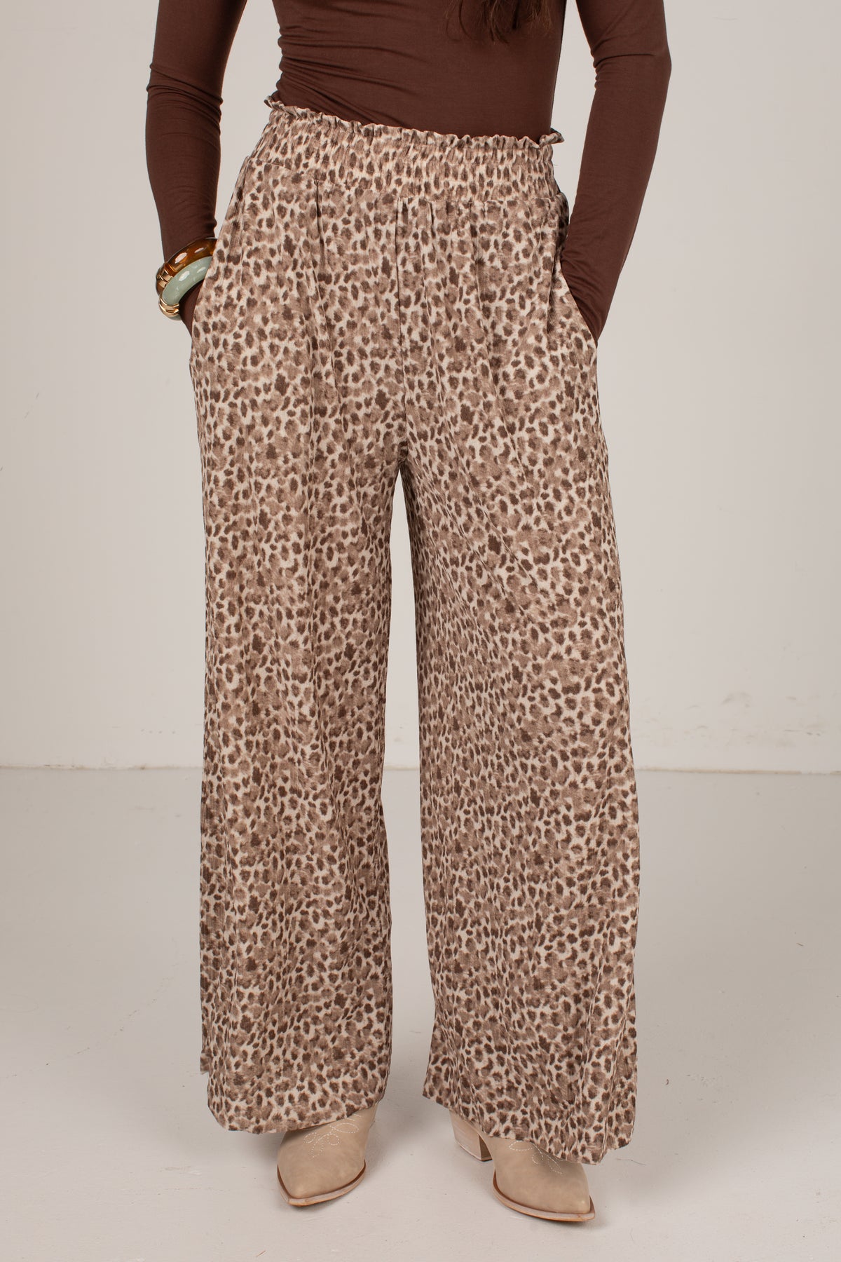 A person wearing high-waisted, wide-leg leopard print pants in beige and brown, paired with a brown long-sleeve top and beige heeled shoes, shown from the waist down against a plain light background.