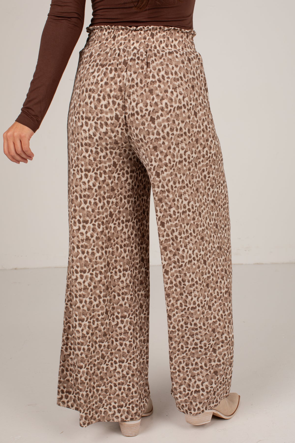 A person wearing high-waisted, wide-leg pants with a brown and beige leopard print pattern, paired with a dark brown long-sleeve top and beige heels, shown from the back against a plain light gray studio background.