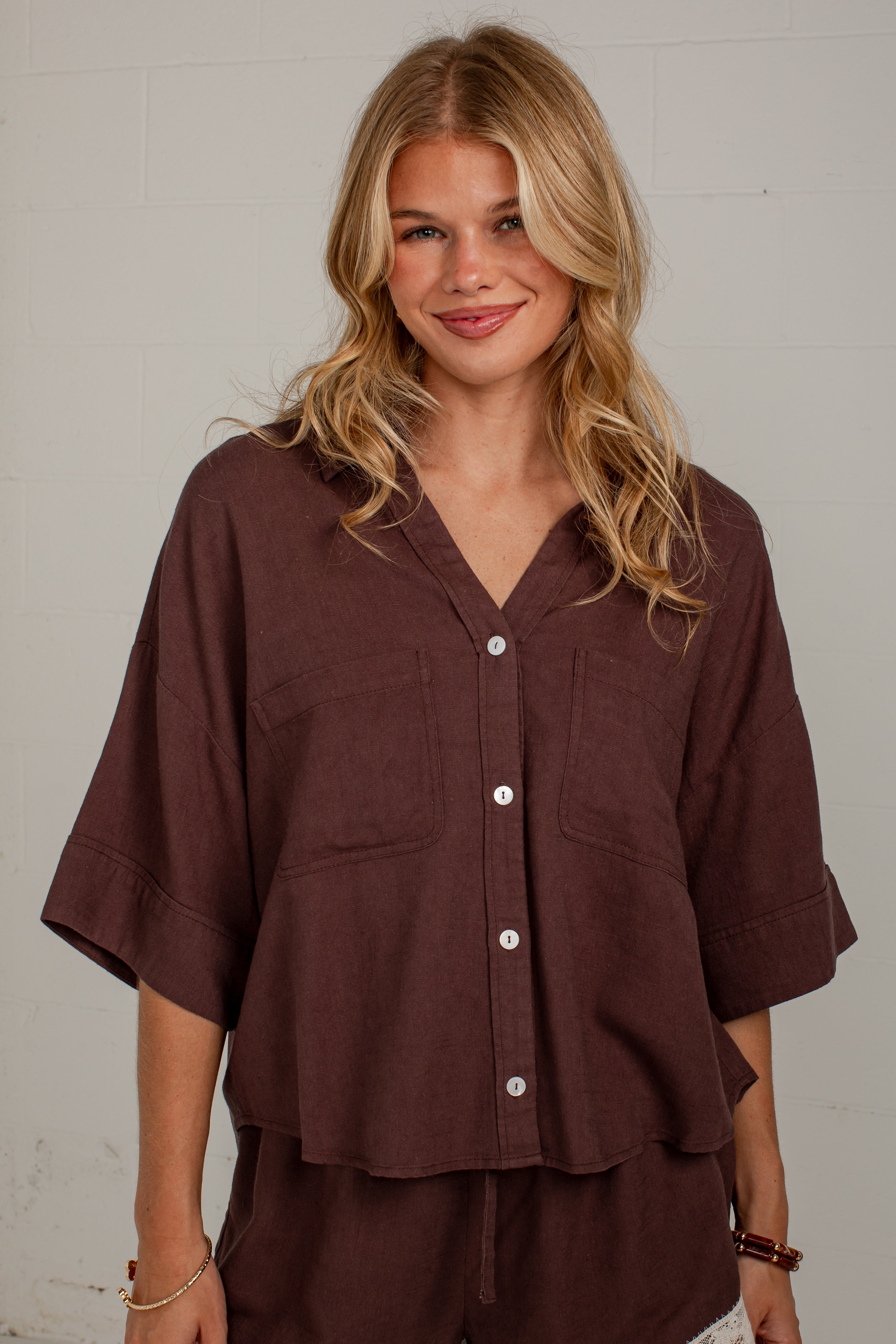 A woman wearing a dark brown short-sleeve button-up shirt with a relaxed, oversized fit and matching brown bottoms, standing against a light gray brick wall.