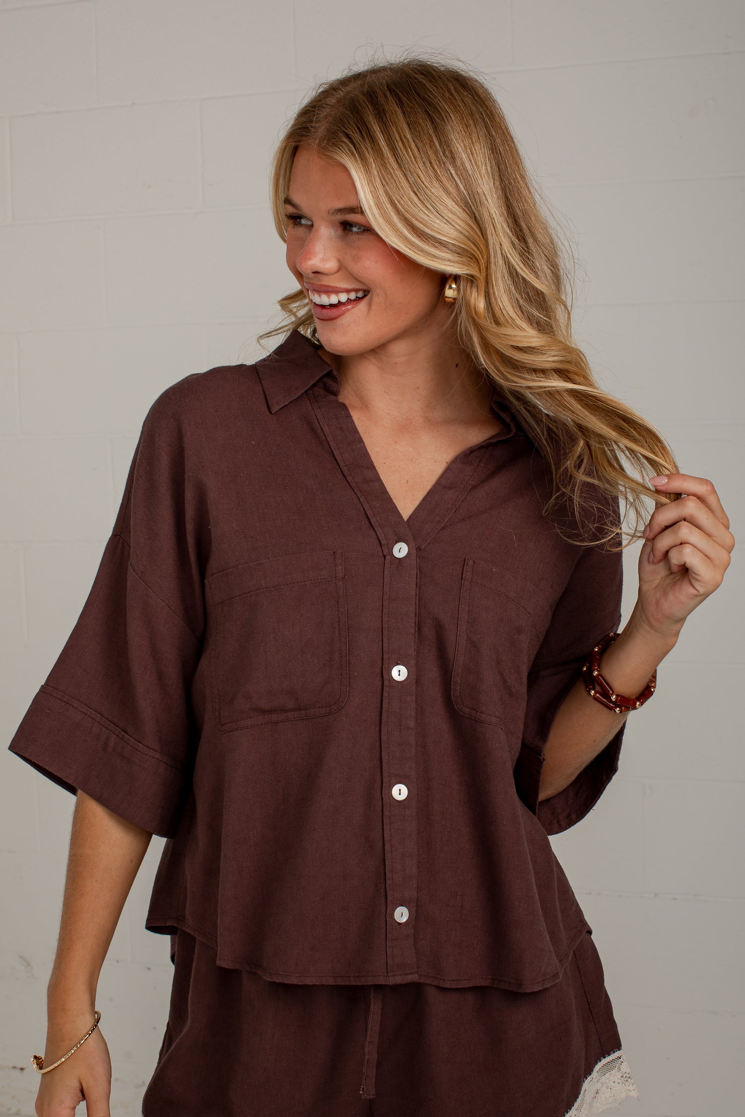 A woman wearing a dark brown short-sleeve button-up shirt with a relaxed fit, shown from the waist up against a light gray wall.