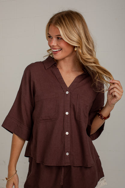 A woman wearing a dark brown short-sleeve button-up shirt with a relaxed fit, shown from the waist up against a light gray wall.