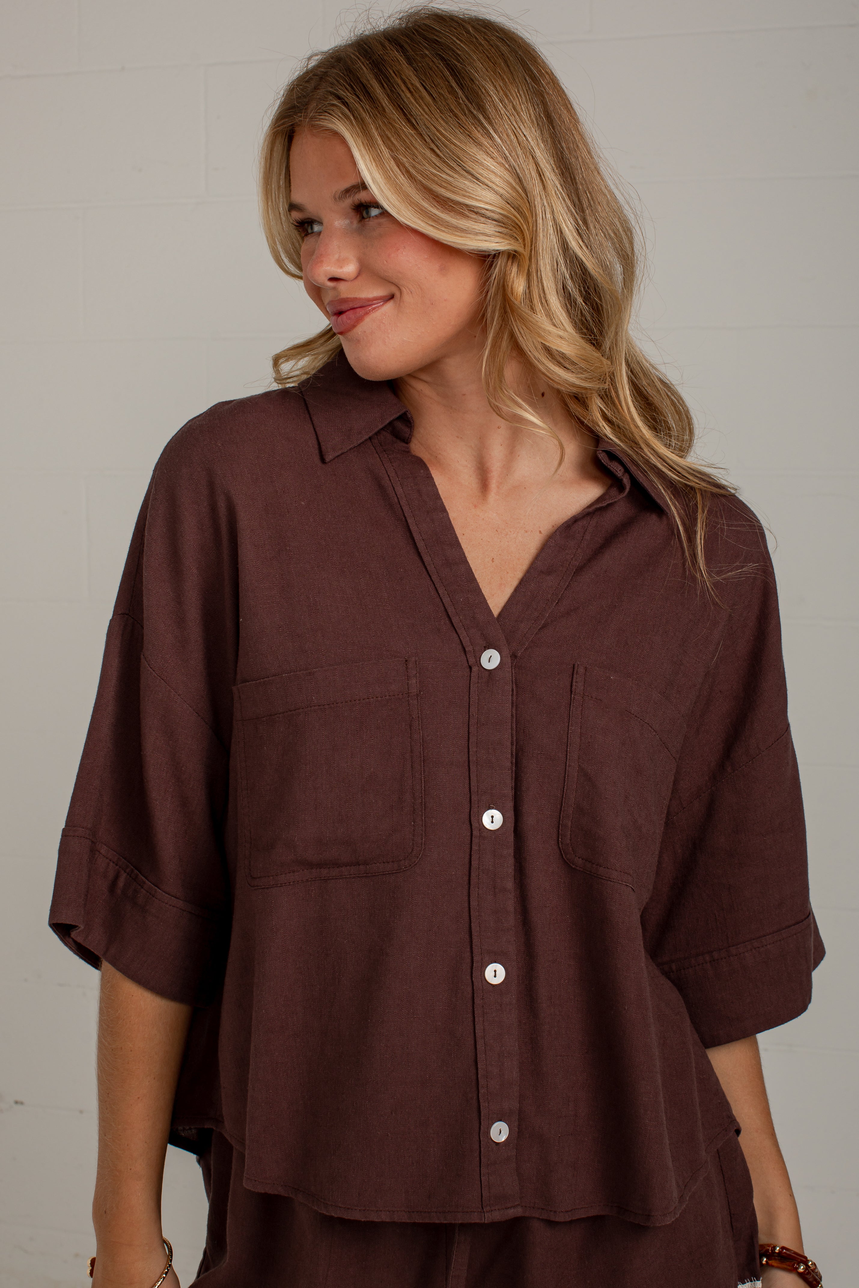 A woman wearing a dark brown button-down shirt with three-quarter sleeves and two chest pockets, photographed against a light neutral wall.