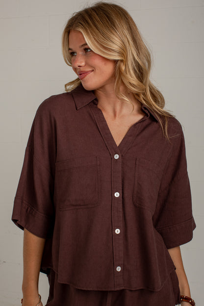 A woman wearing a dark brown button-down shirt with three-quarter sleeves and two chest pockets, photographed against a light neutral wall.