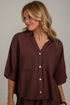 A woman wearing a dark brown button-down shirt with three-quarter sleeves and two chest pockets, photographed against a light neutral wall.