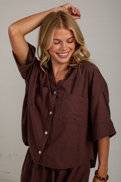A woman wearing a dark brown short-sleeve button-up shirt with a chest pocket, posing against a light gray wall.