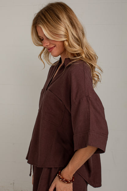 A woman with blonde hair wearing a dark brown linen-like short-sleeve top and matching pants, photographed against a light off-white wall.