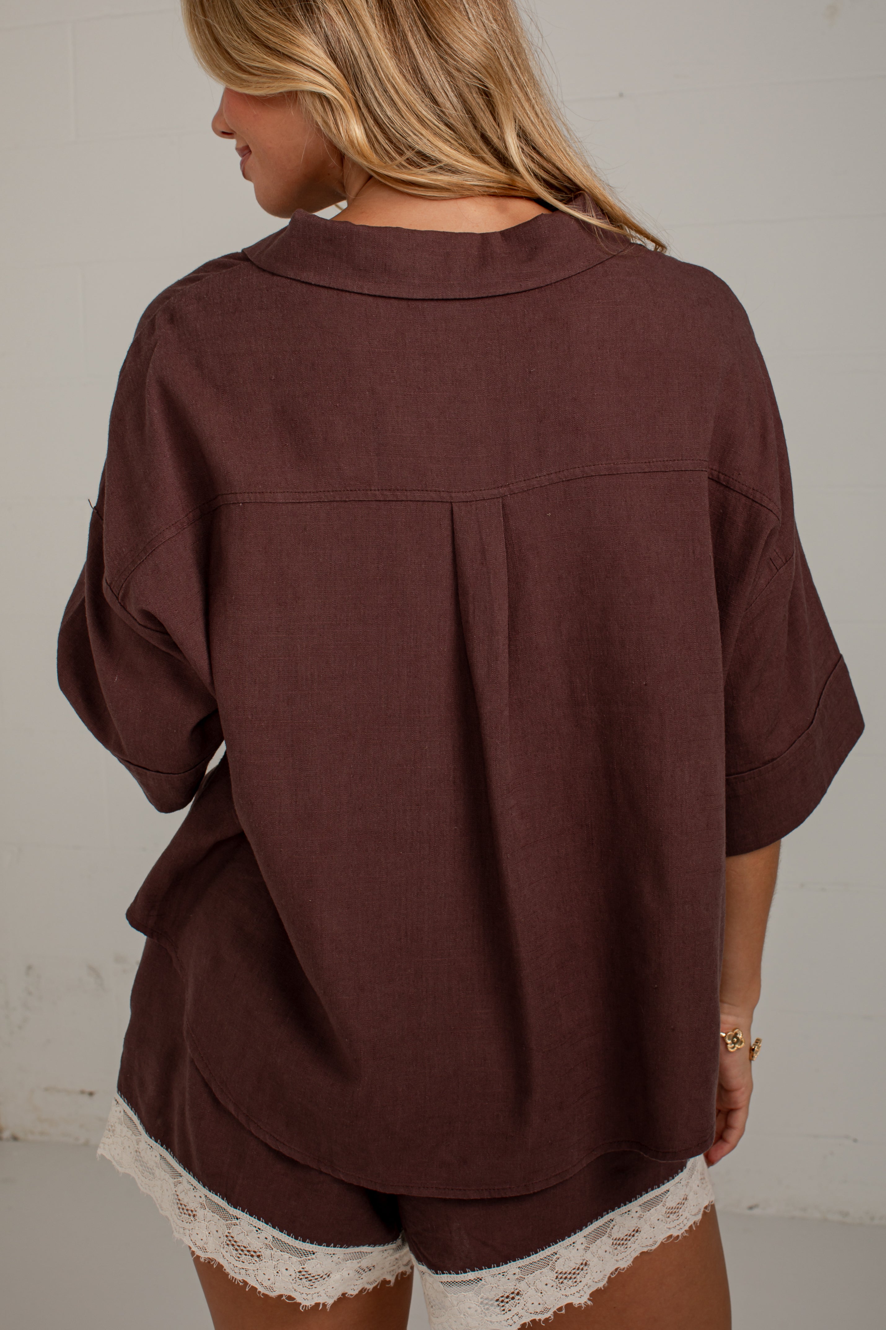 A woman viewed from behind wearing a loose-fitting dark brown short-sleeve top with a pleated back detail and matching shorts featuring white lace trim, set against a plain off-white wall.