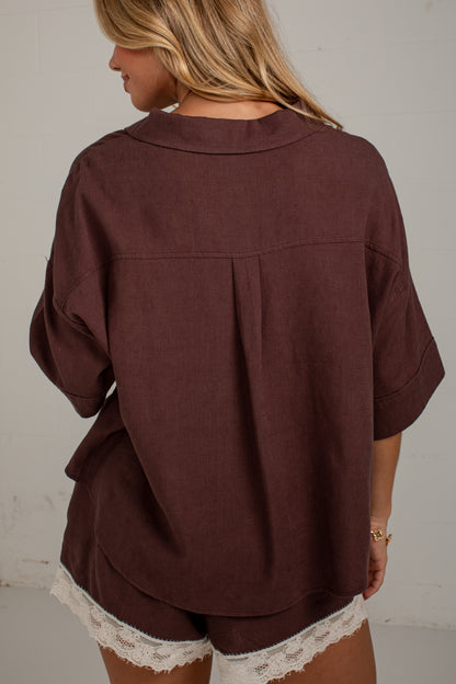 A woman viewed from behind wearing a loose-fitting dark brown short-sleeve top with a pleated back detail and matching shorts featuring white lace trim, set against a plain off-white wall.