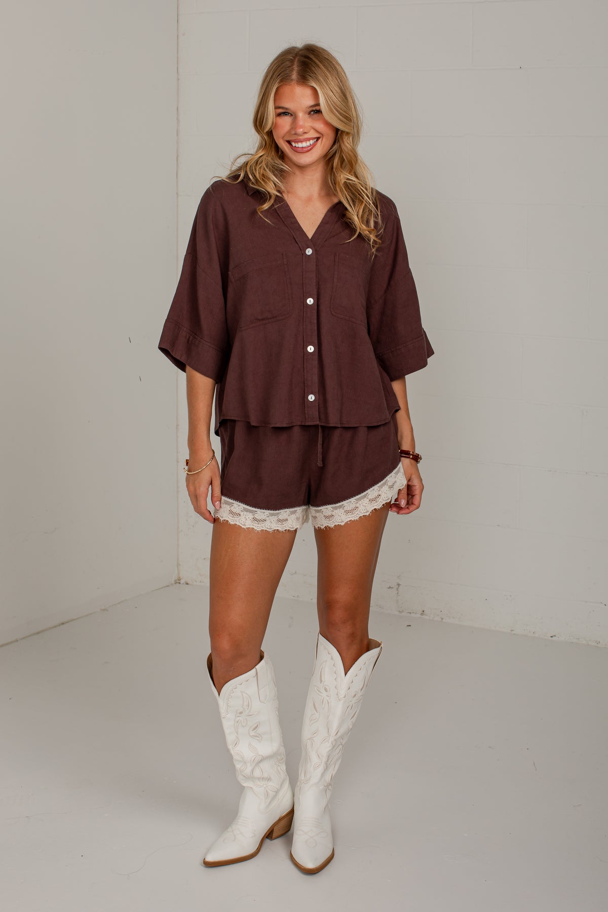 A woman modeling a matching brown short-sleeve button-front top and shorts set with white lace trim, paired with white cowboy boots, in a plain studio setting.