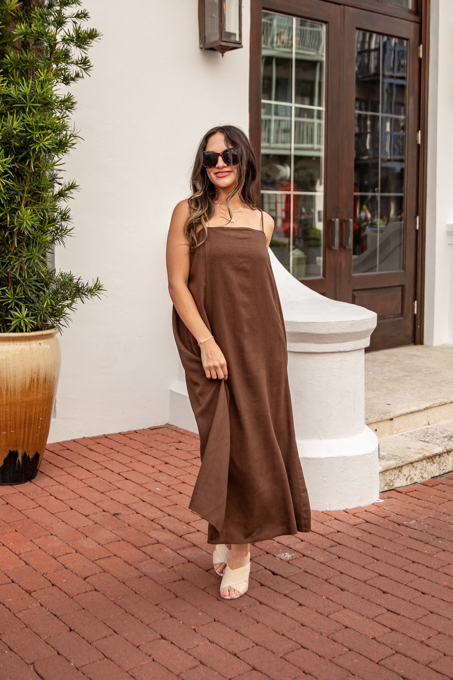 Woman in a brown dress standing on a brick patio with a plant and building entrance in the background.