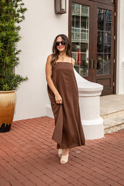Woman in a brown dress standing on a brick patio with a plant and building entrance in the background.