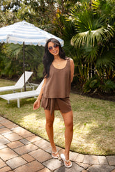 Woman in a brown outfit standing in a garden with palm trees and lounge chairs.