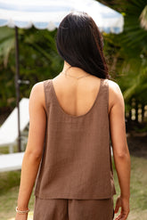 Woman wearing a brown sleeveless top and pants in an outdoor setting with greenery.