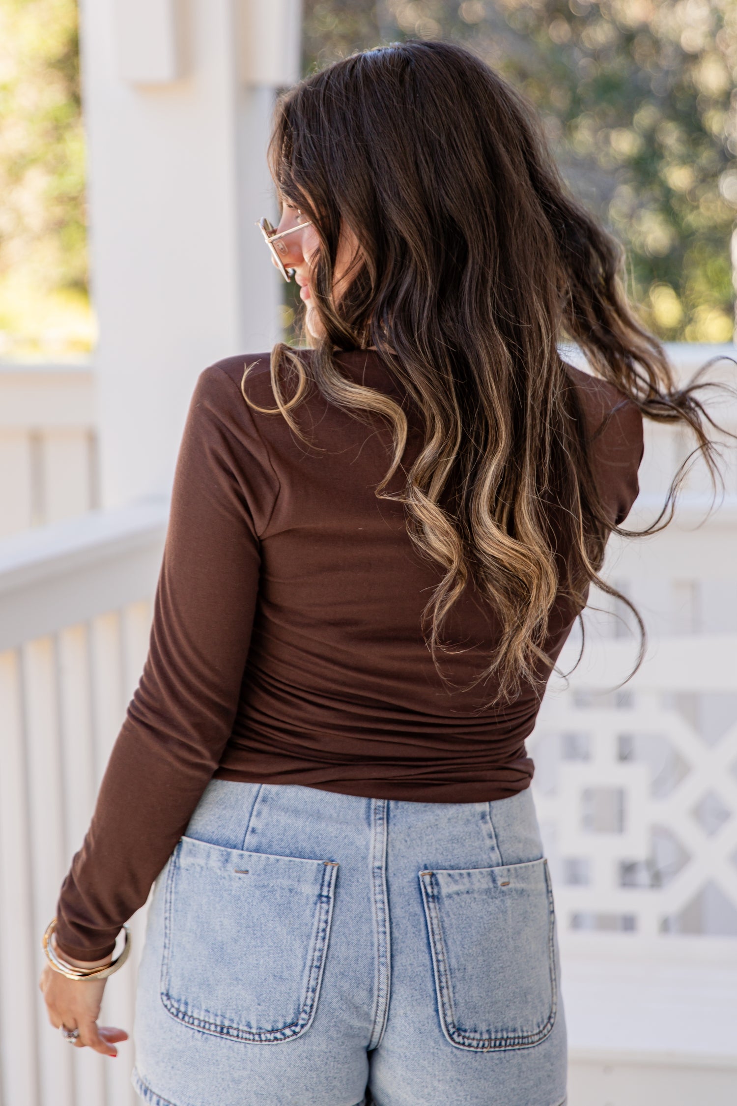 Woman wearing a brown long-sleeve top and light blue jeans on a balcony.