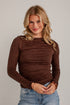 A woman with blonde hair wearing a brown long-sleeve ruched top and blue jeans, standing against a white brick wall.