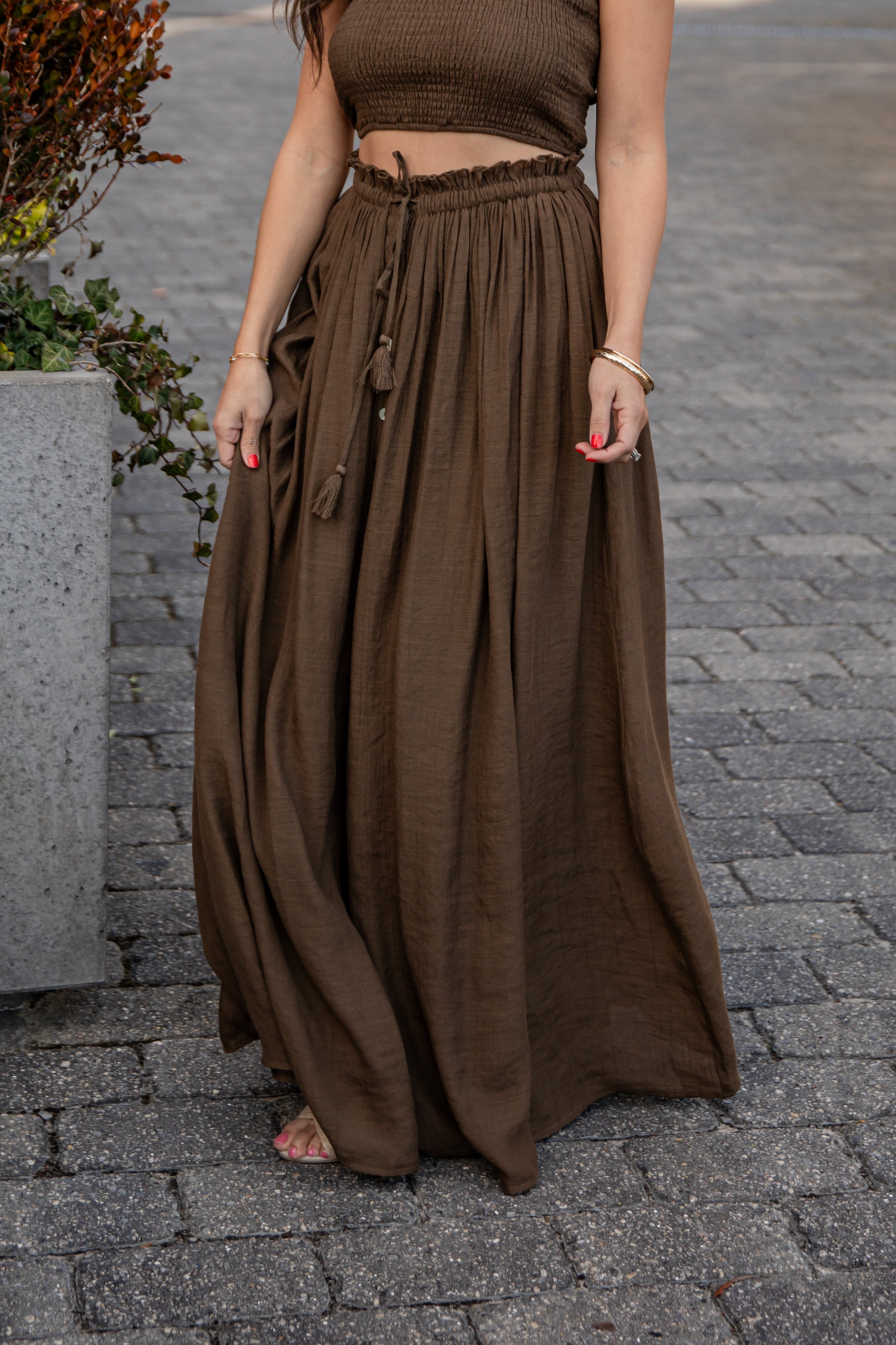 Woman wearing a brown top and long skirt standing on a paved street.