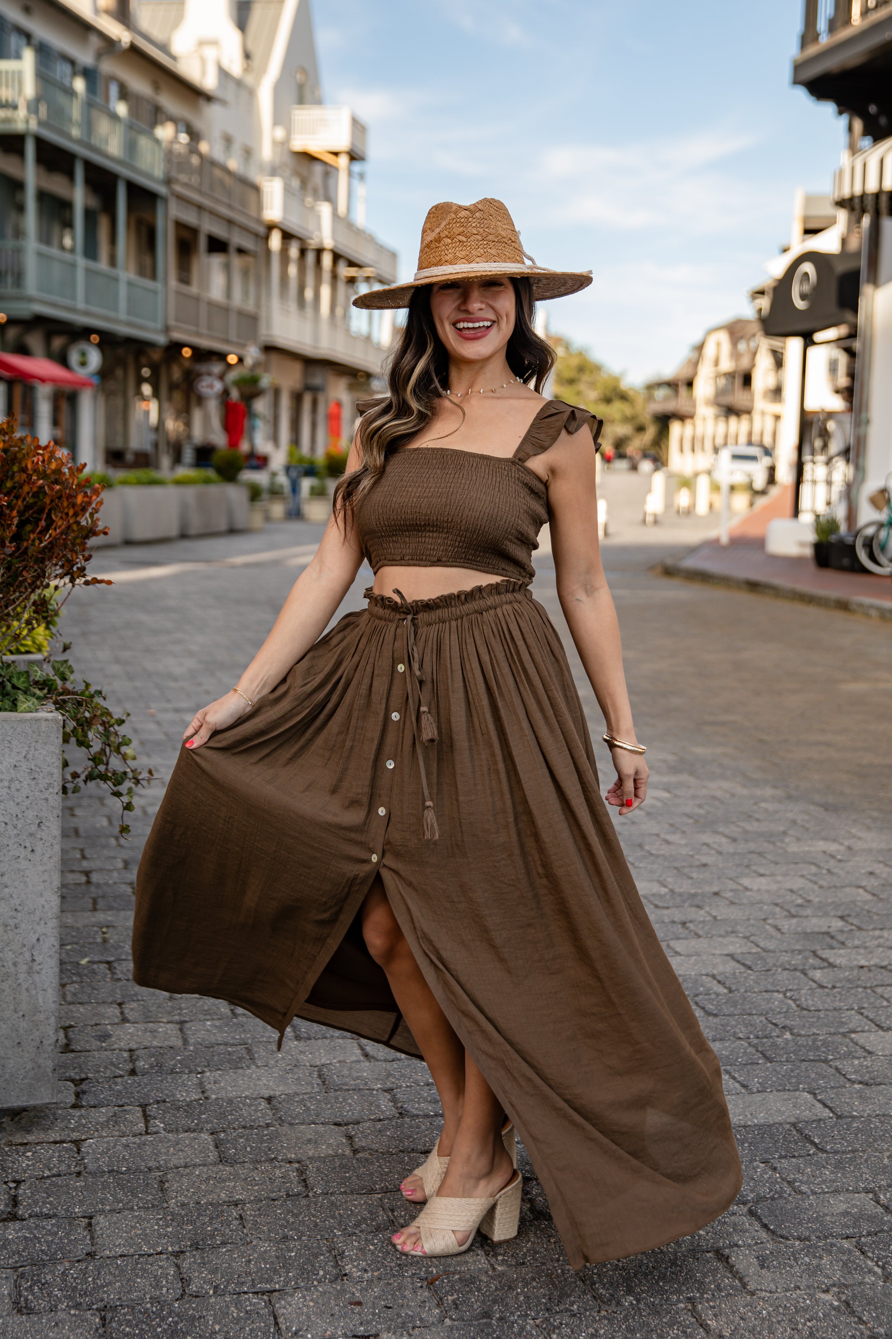 Woman in a brown dress and hat standing on a street with buildings and trees in the background