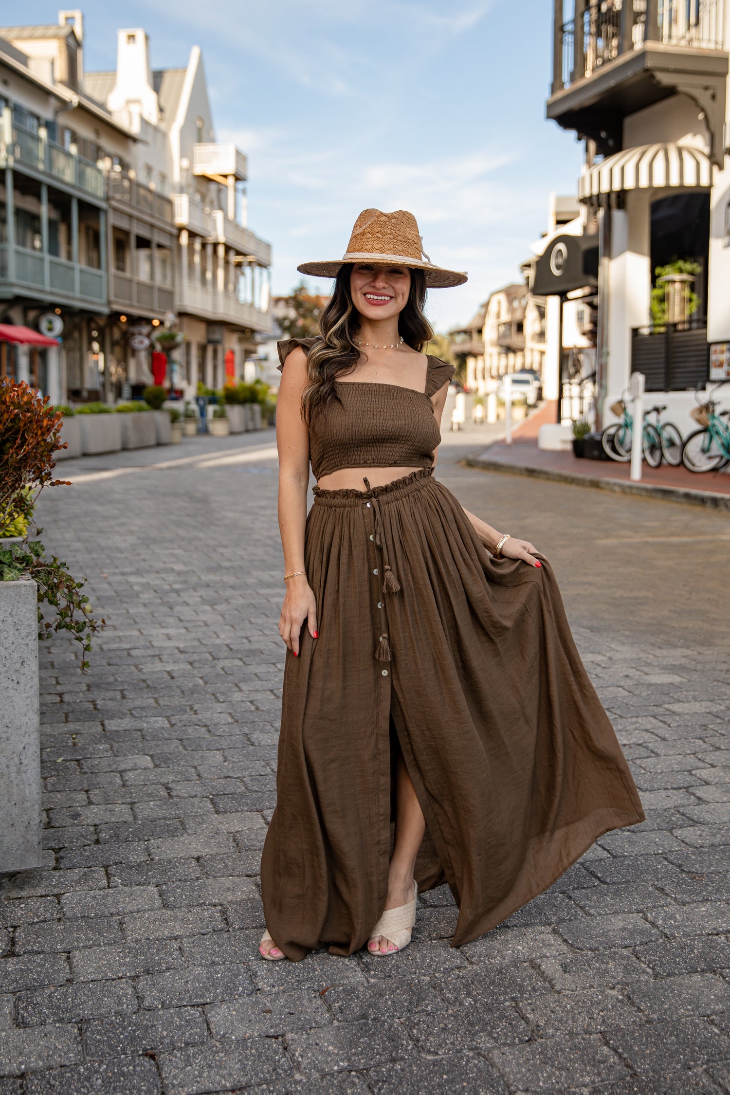 Woman in a brown dress and hat standing on a street with buildings and bicycles in the background.