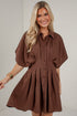 A smiling blonde woman wearing a brown short-sleeve button-front shirt dress with a flared skirt, standing against a light gray wall.