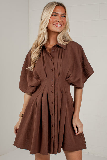 A woman wearing a brown short-sleeve button-up shirt dress with a flared skirt, standing against a white brick wall.