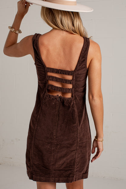 A woman shown from the back wearing a dark brown corduroy sleeveless dress with a ruffled cutout back, paired with a wide-brimmed light beige hat, against a white brick wall.