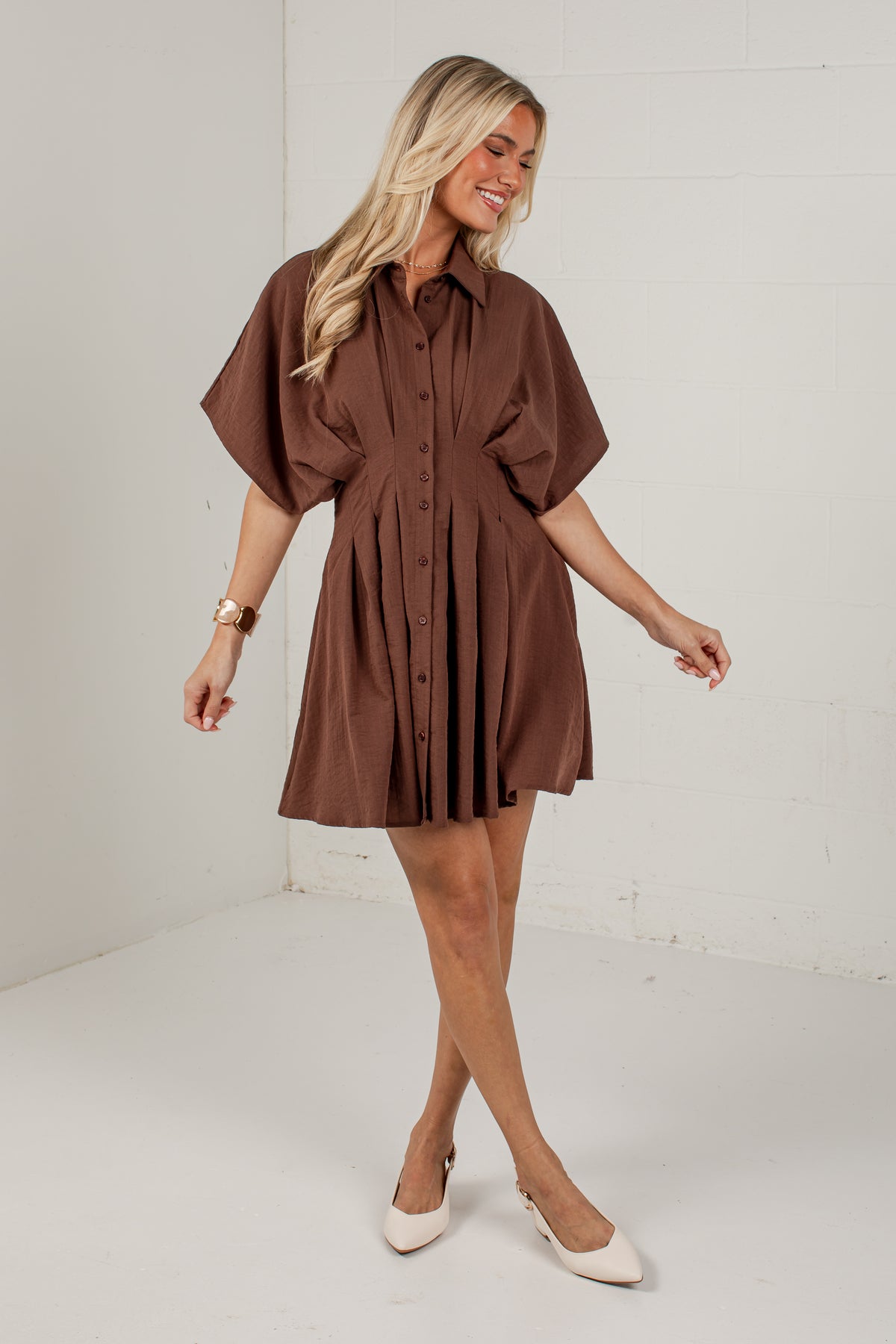 A woman modeling a brown short-sleeve button-front shirt dress with a pleated skirt, standing in a bright white studio setting.