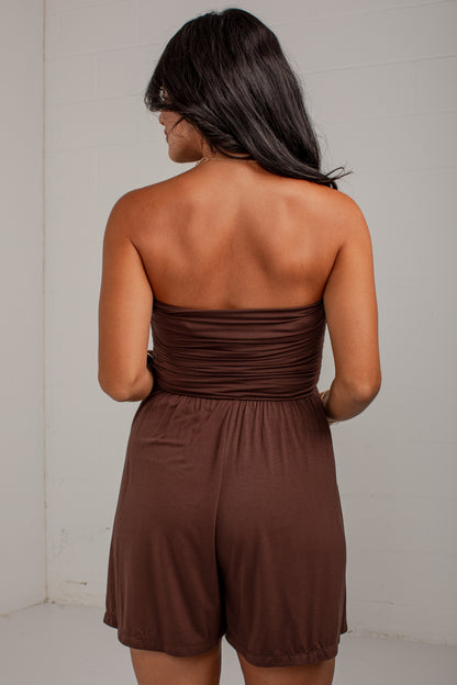 A woman shown from the back wearing a brown strapless romper with a ruched waistband and short legs, standing against a plain light wall.