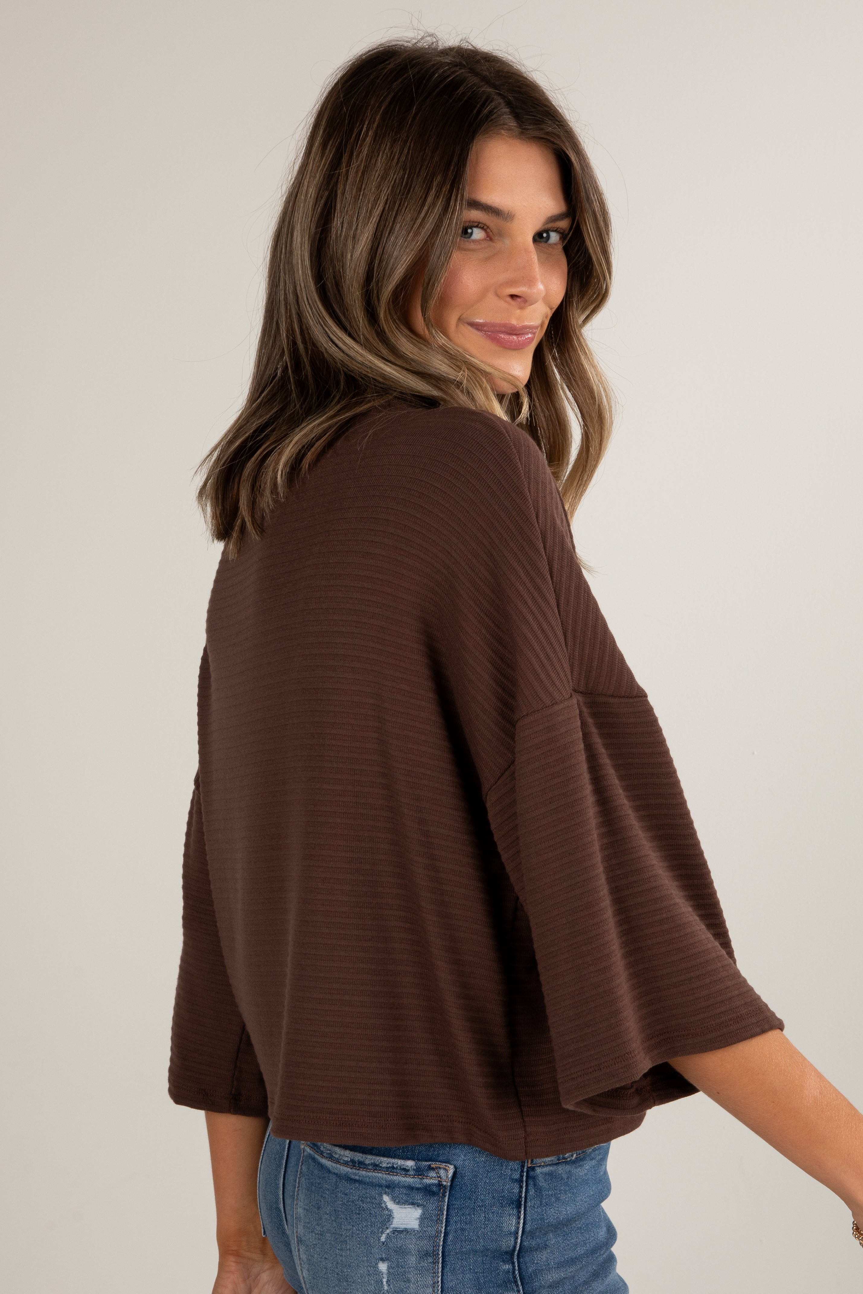 Woman wearing a brown ribbed sweater and blue jeans against a plain background