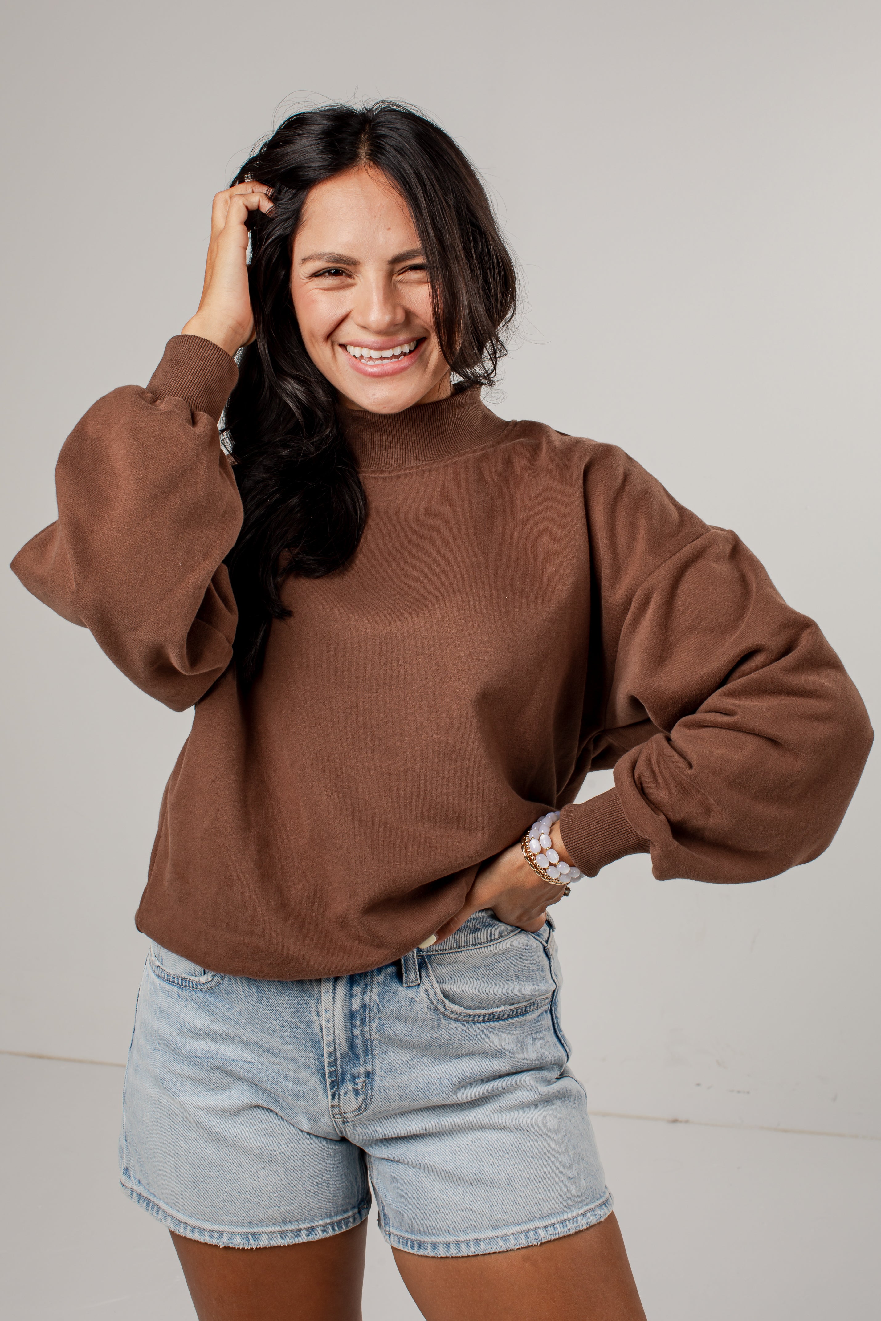 A woman smiling and posing in a brown mock-neck sweatshirt with voluminous balloon sleeves, paired with light-wash denim shorts, against a plain light gray studio background.