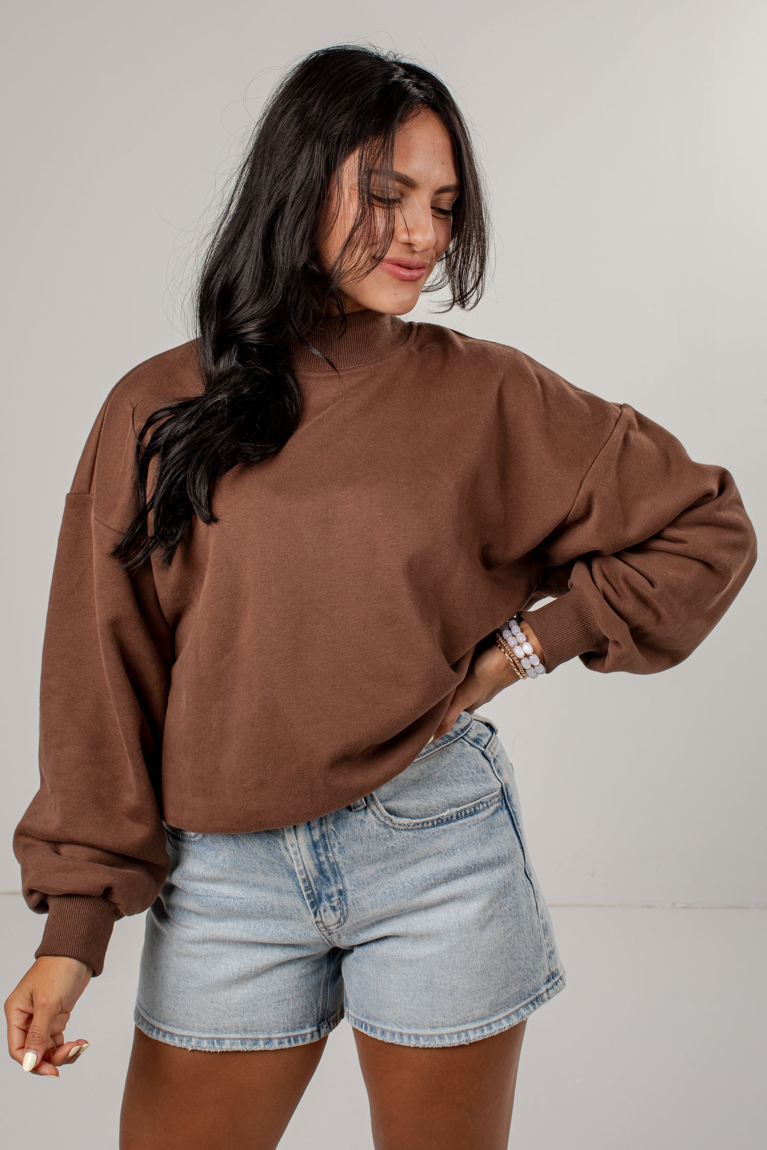 A woman wearing a brown long-sleeve mock neck sweatshirt and light-wash denim shorts, standing against a neutral light gray studio background.