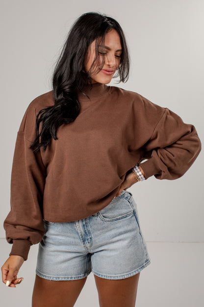 A woman wearing a brown long-sleeve mock neck sweatshirt and light-wash denim shorts, standing against a neutral light gray studio background.