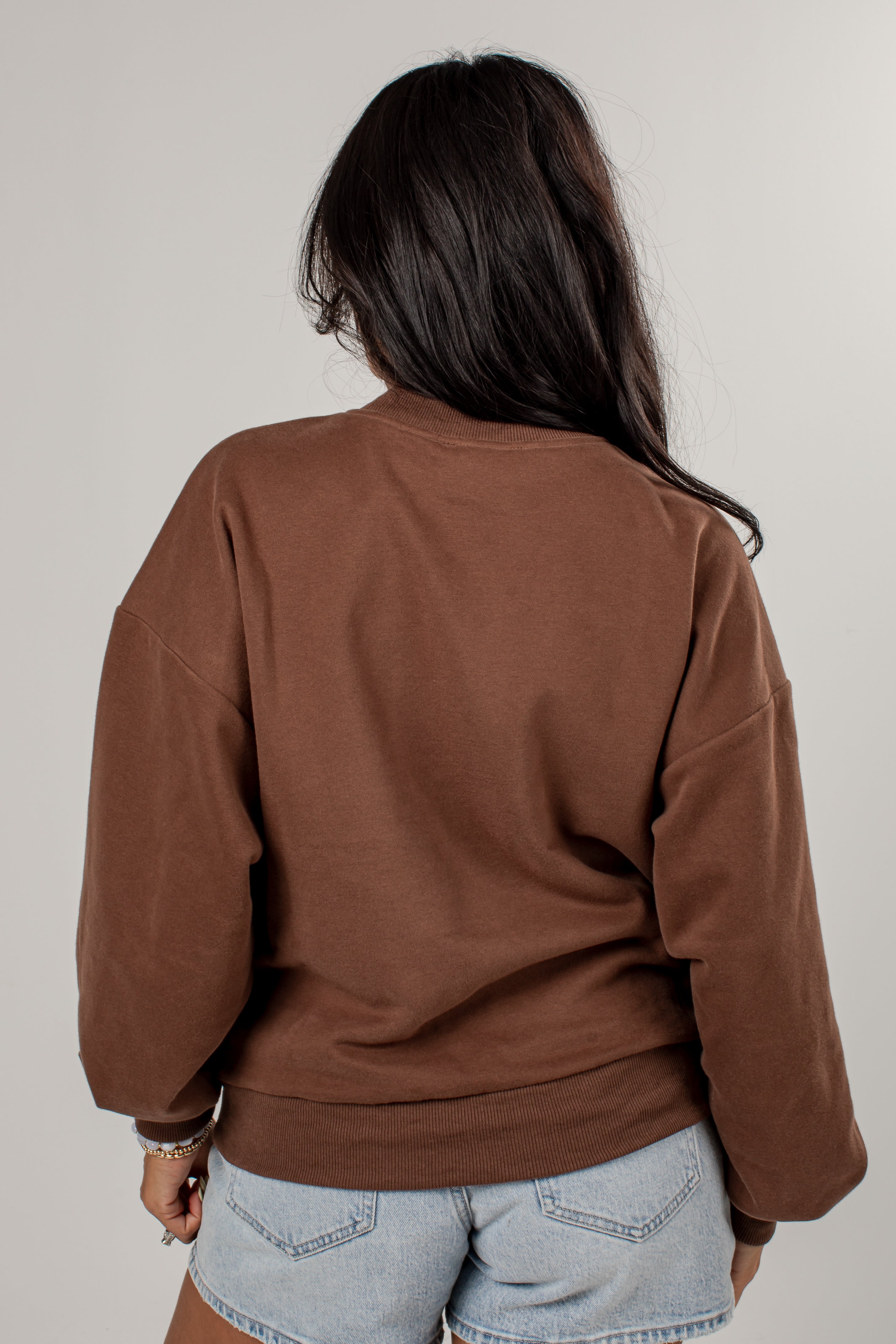 A woman viewed from behind wearing a brown crewneck sweatshirt and light blue denim shorts against a plain light gray background.