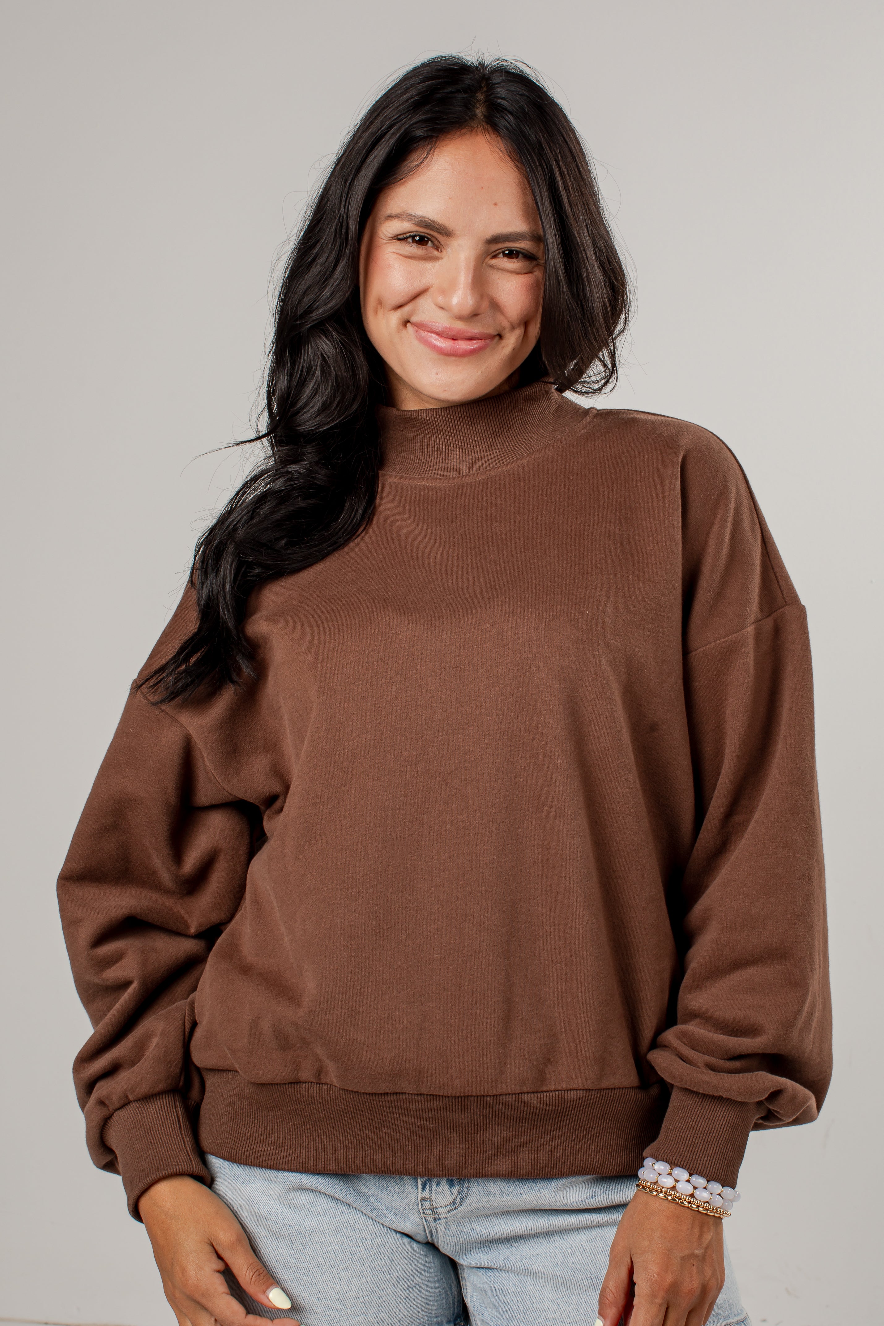 A woman wearing a brown crewneck sweatshirt and light blue jeans, standing against a solid light gray background.